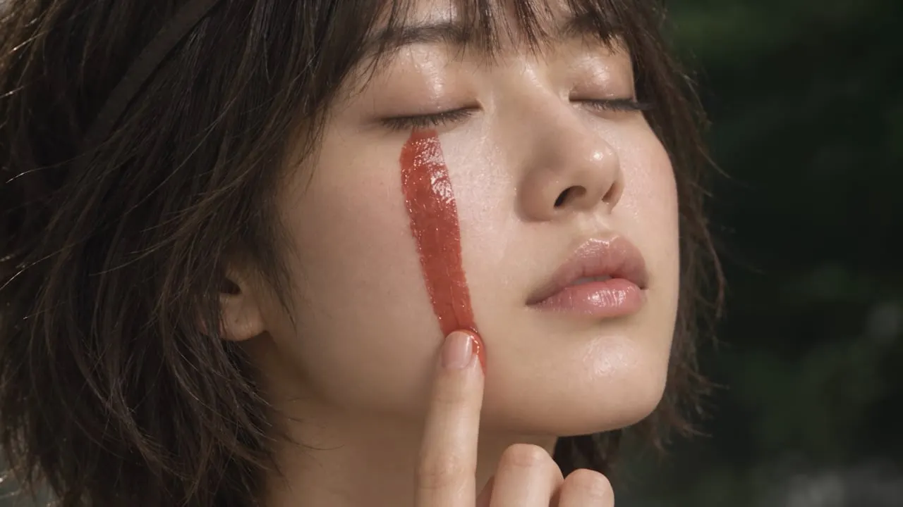 A woman with her eyes closed draws a line of red cream down her cheek.